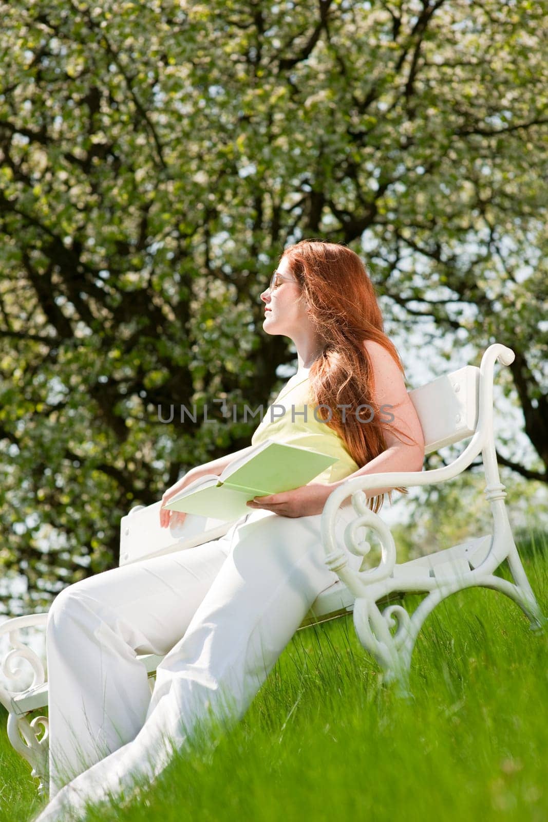 Red hair woman sitting on white bench in green meadow, shallow DOF