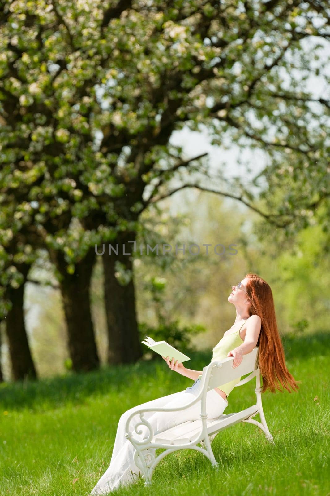 Red hair woman enjoying sun on white bench in green meadow, shallow DOF