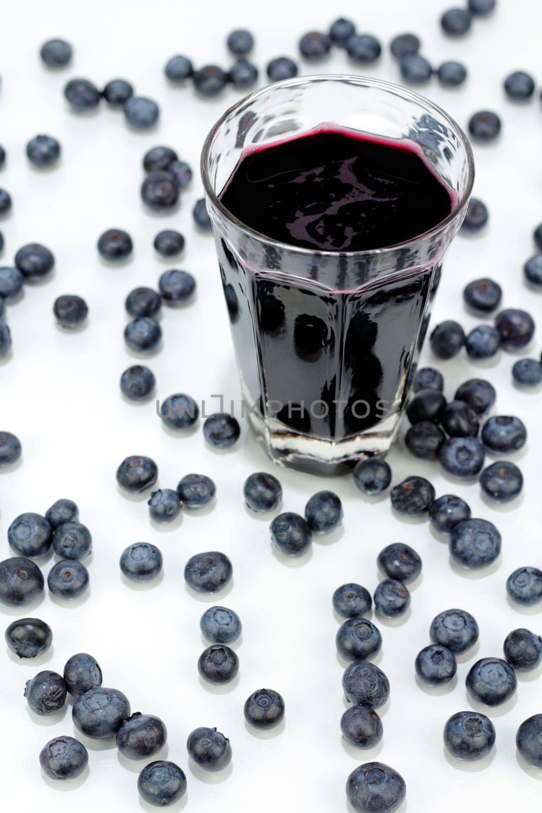 Deep purple berry juice in clear glass, whole fruit abstract on white reflective surface.