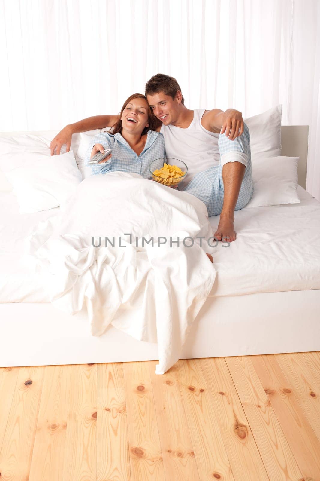 Young man and woman lying down in white bed watching television and eating crisps