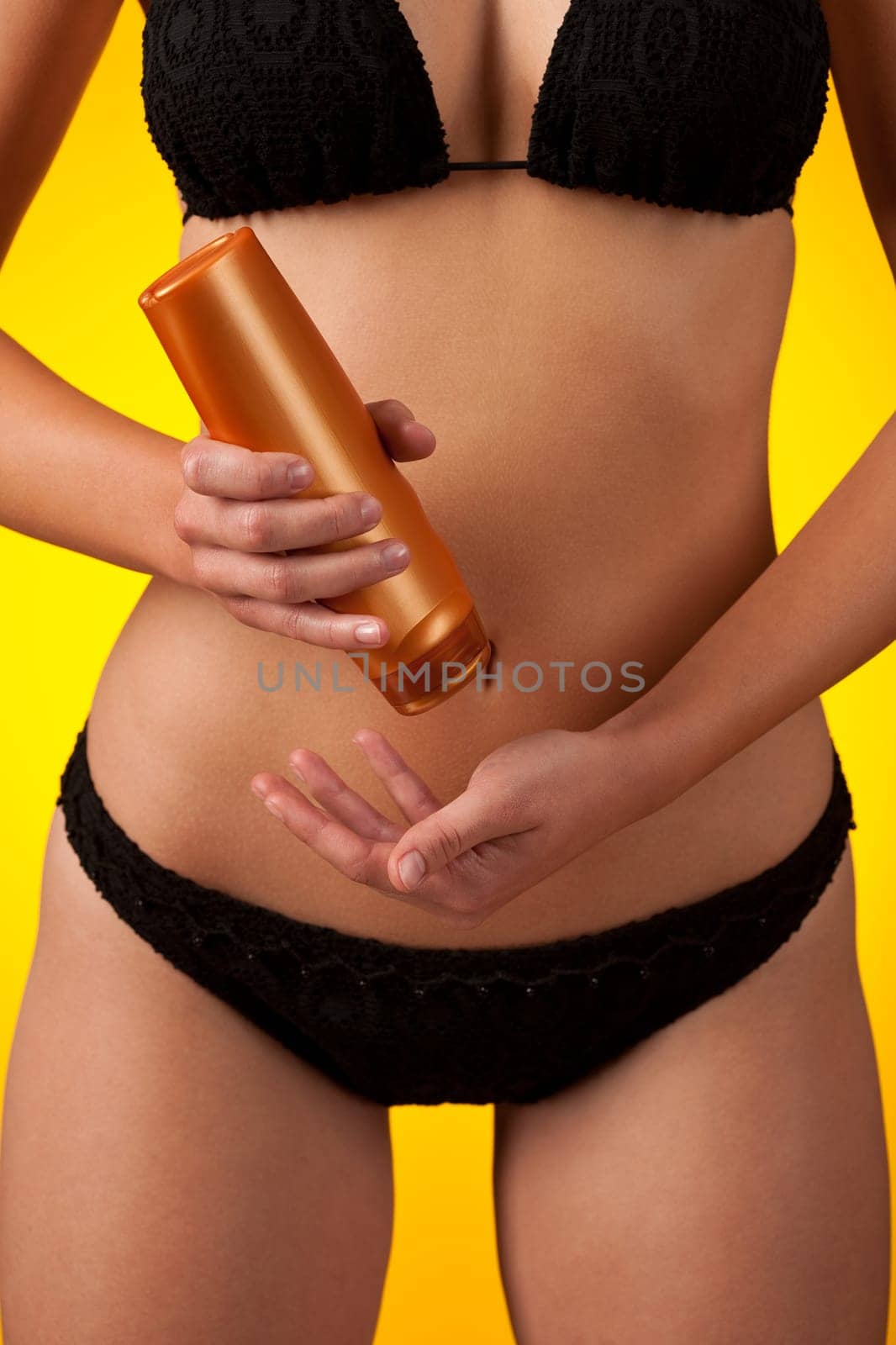 Part of female body wearing black bikini and bottle of suntan cream on yellow background