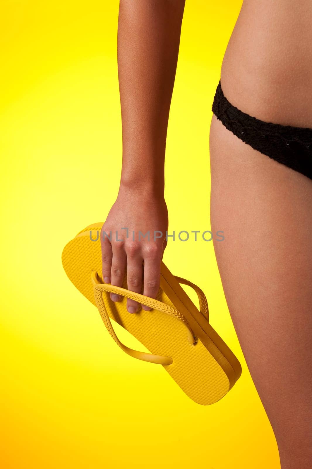 Part of female body wearing black bikini and holding flip-flop on yellow background
