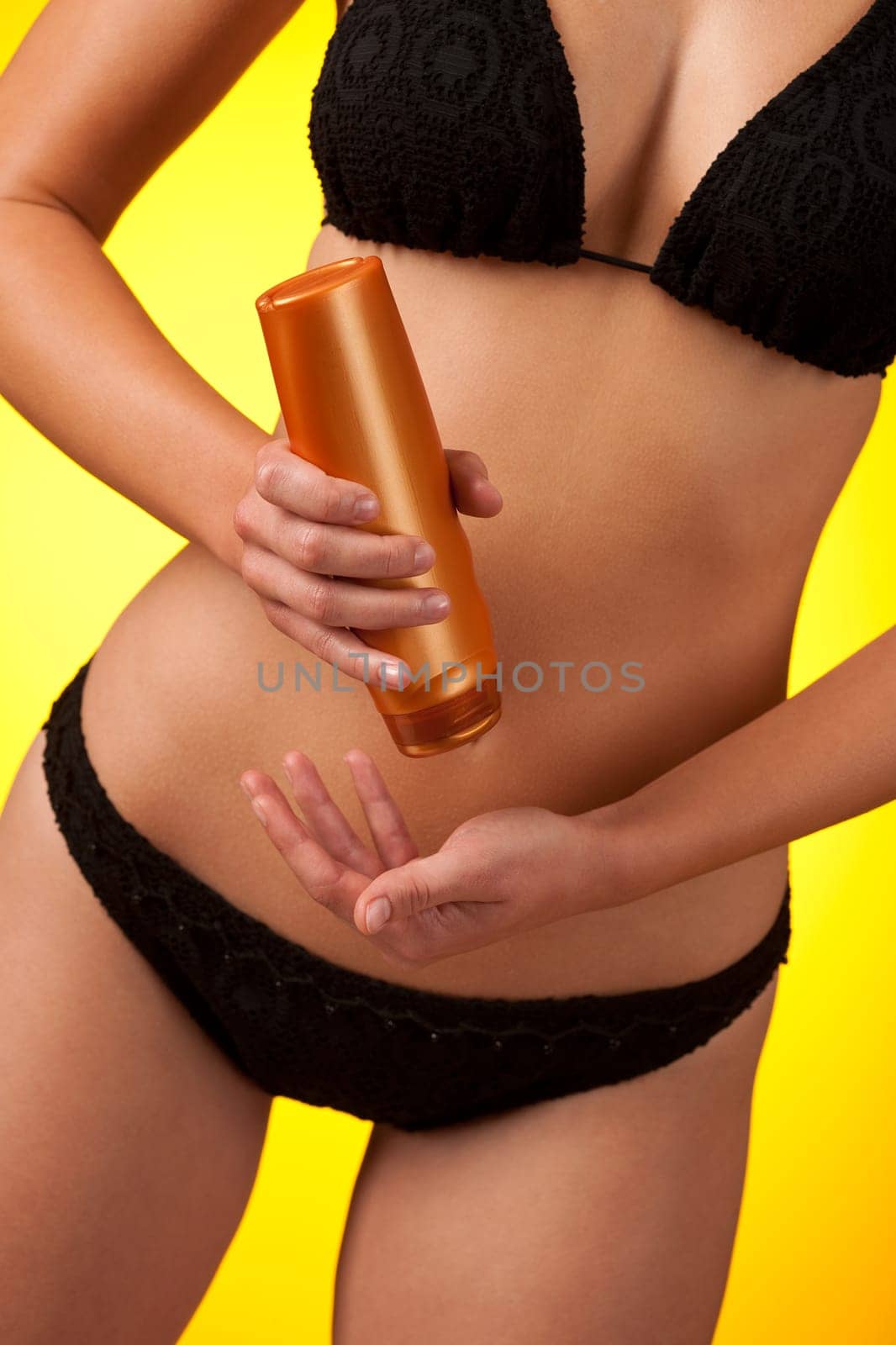 Part of female body wearing black bikini and bottle of suntan cream on yellow background
