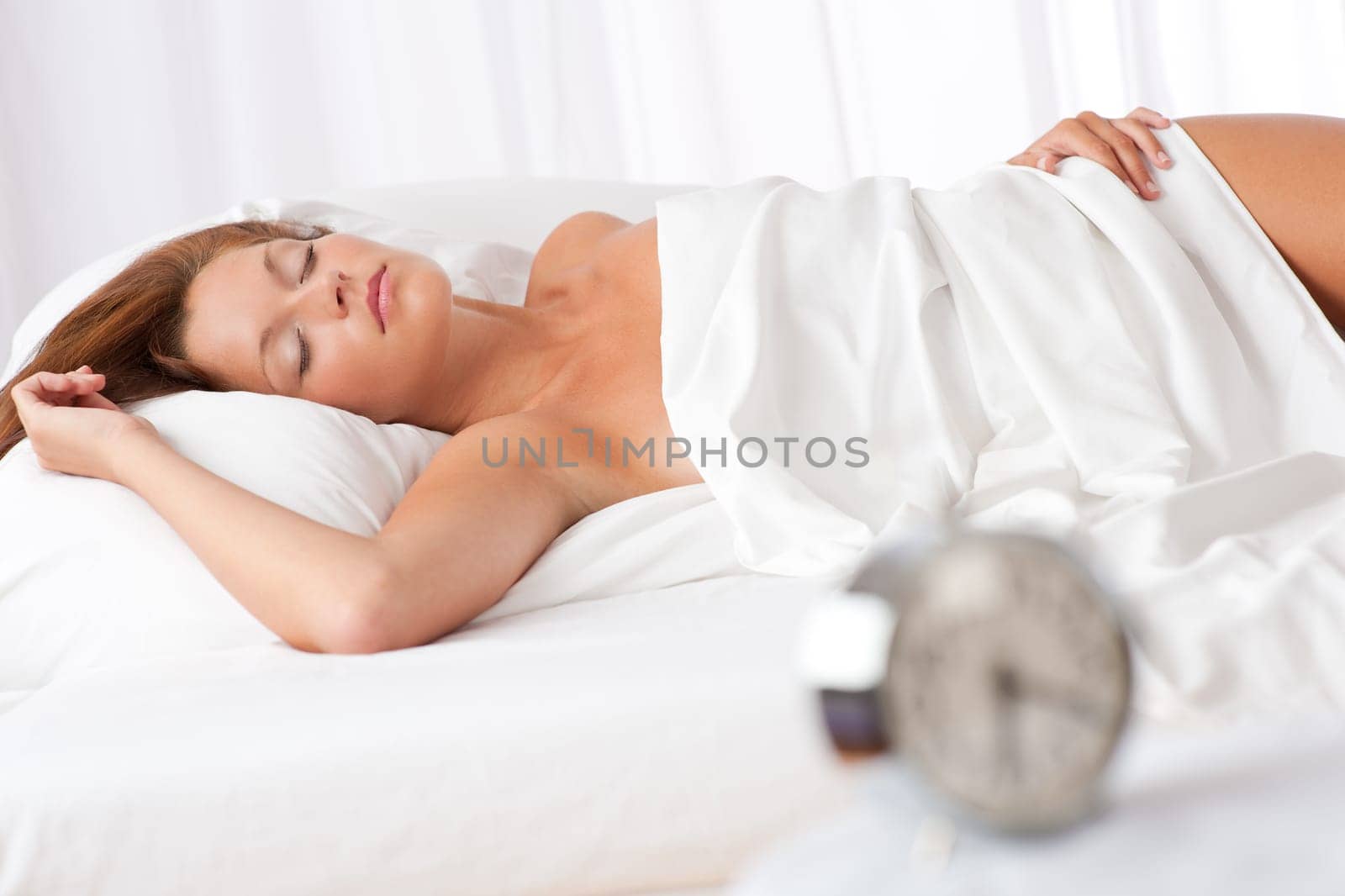 Woman sleeping in white bed, alarm clock in foreground