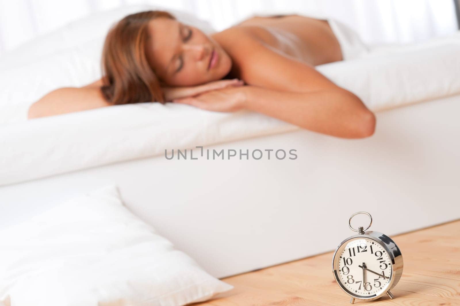 Young woman sleeping on white bed, alarm clock in foreground