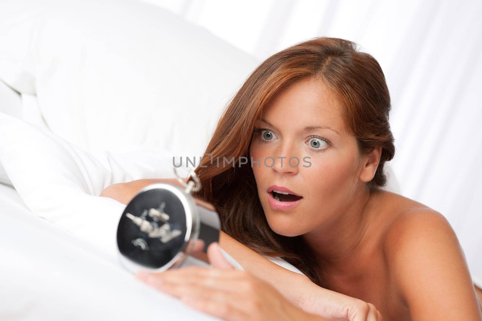Shocked woman with alarm clock lying in white bed