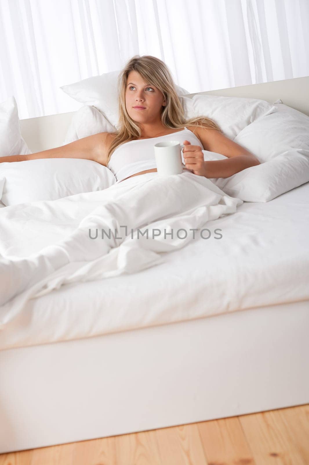Blond woman lying in white bed drinking coffee relaxing