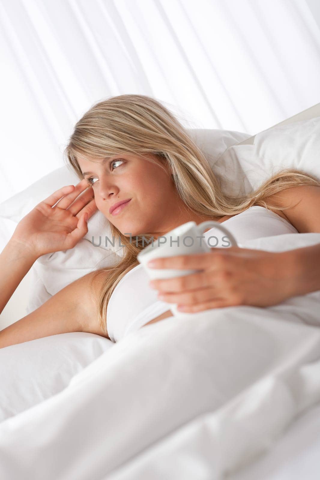 Blond woman lying in white bed holding cup of coffee