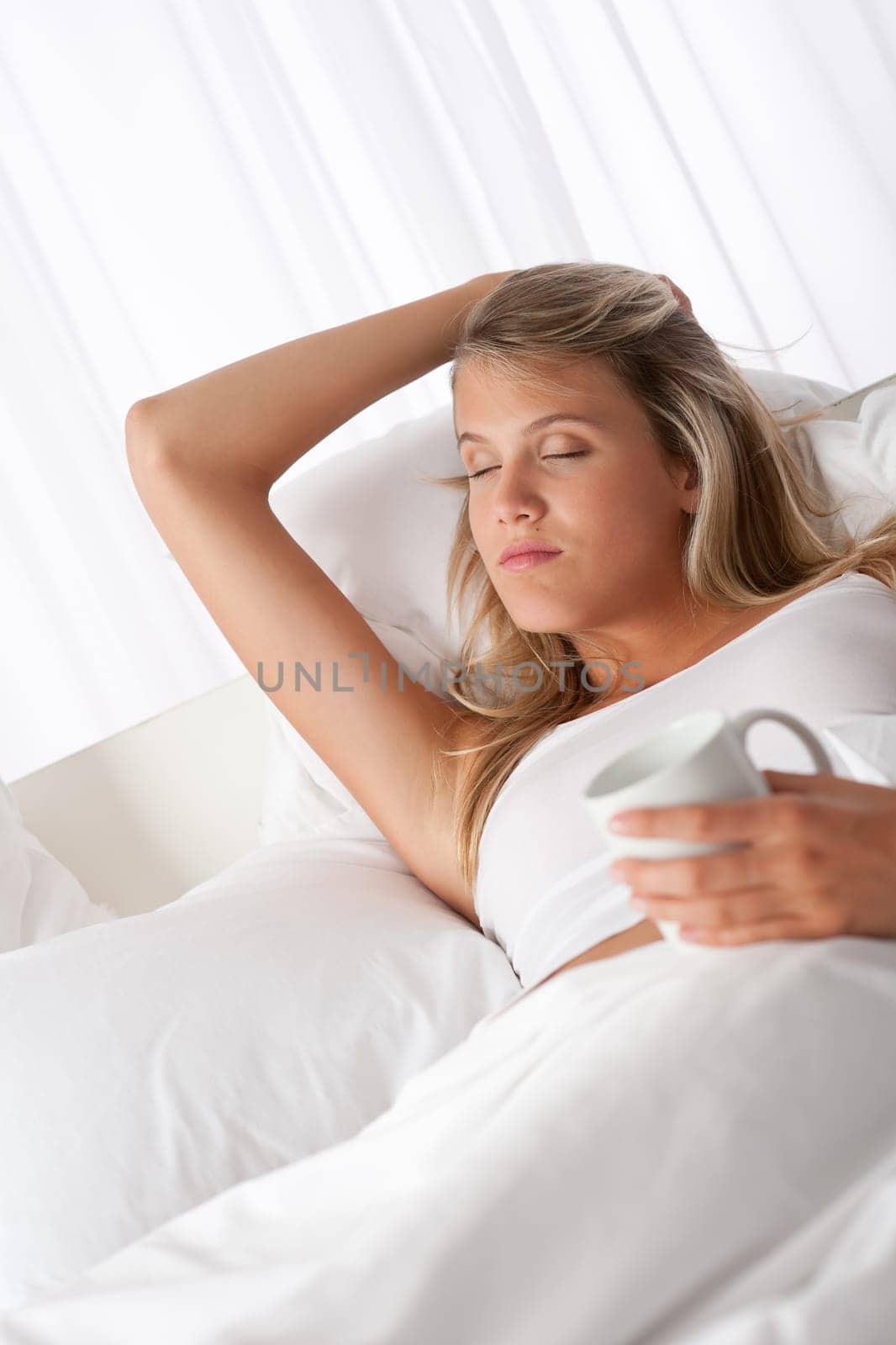 Blond woman holding white mug with coffee lying on white bed