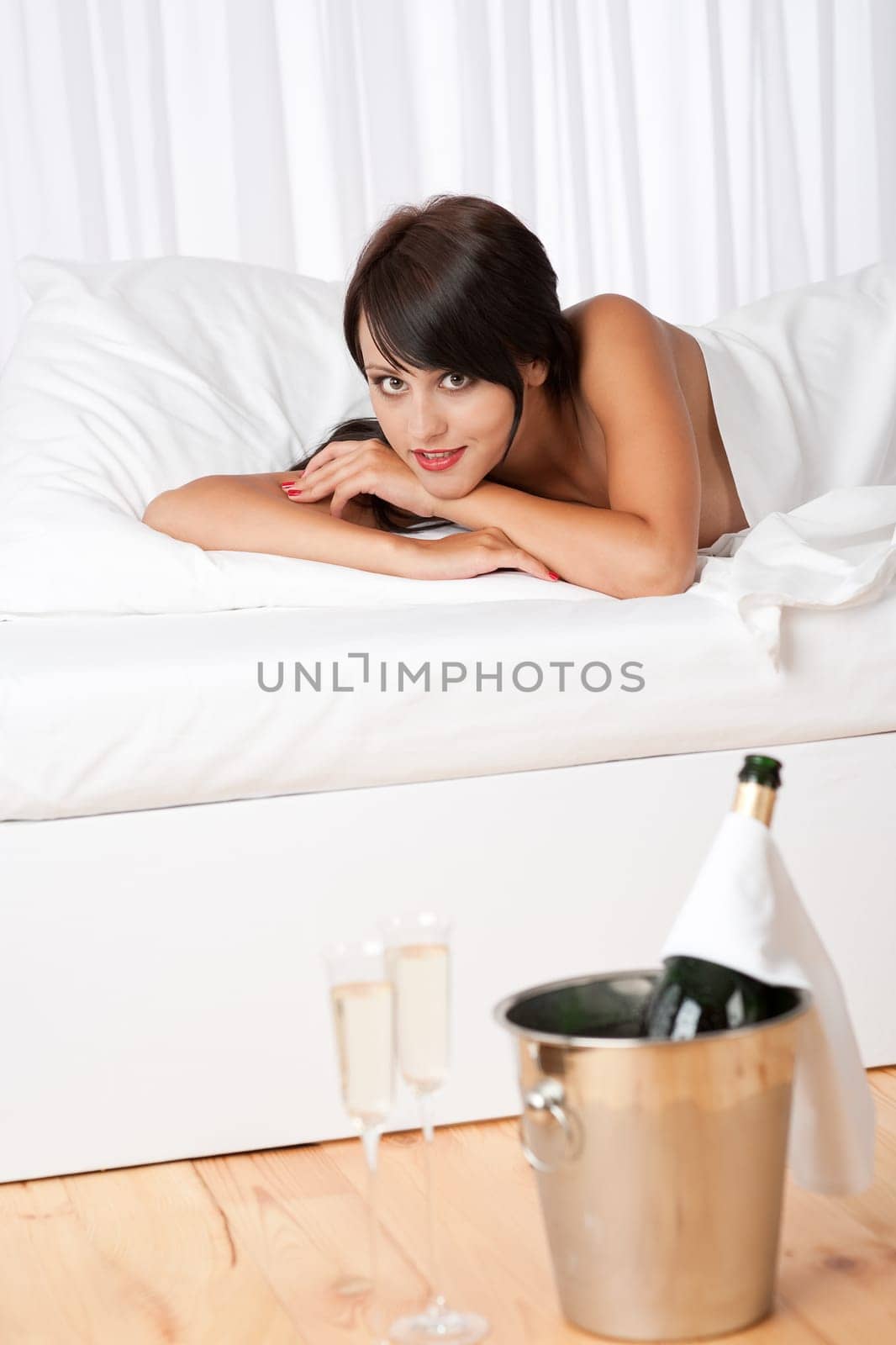 Naked woman lying in white bed with champagne and two glasses