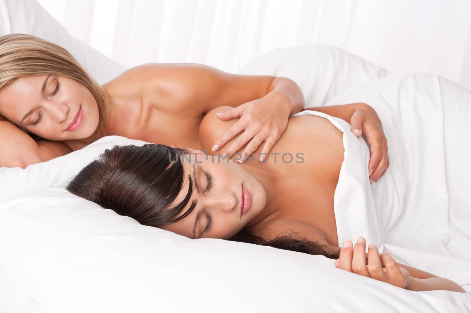 Blond woman and brunette lying down in white bed and sleeping naked together