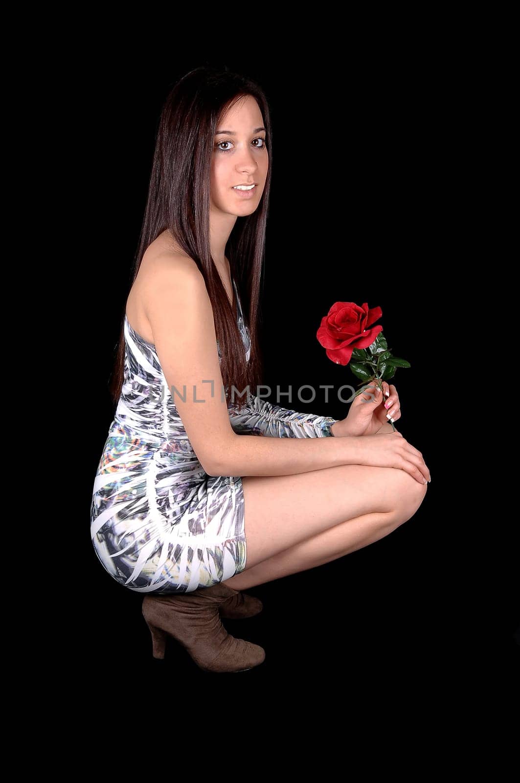 A beautiful young woman in a short tight dress, hocking on the floor in profile in the studio with a rose in her hand, for black background.