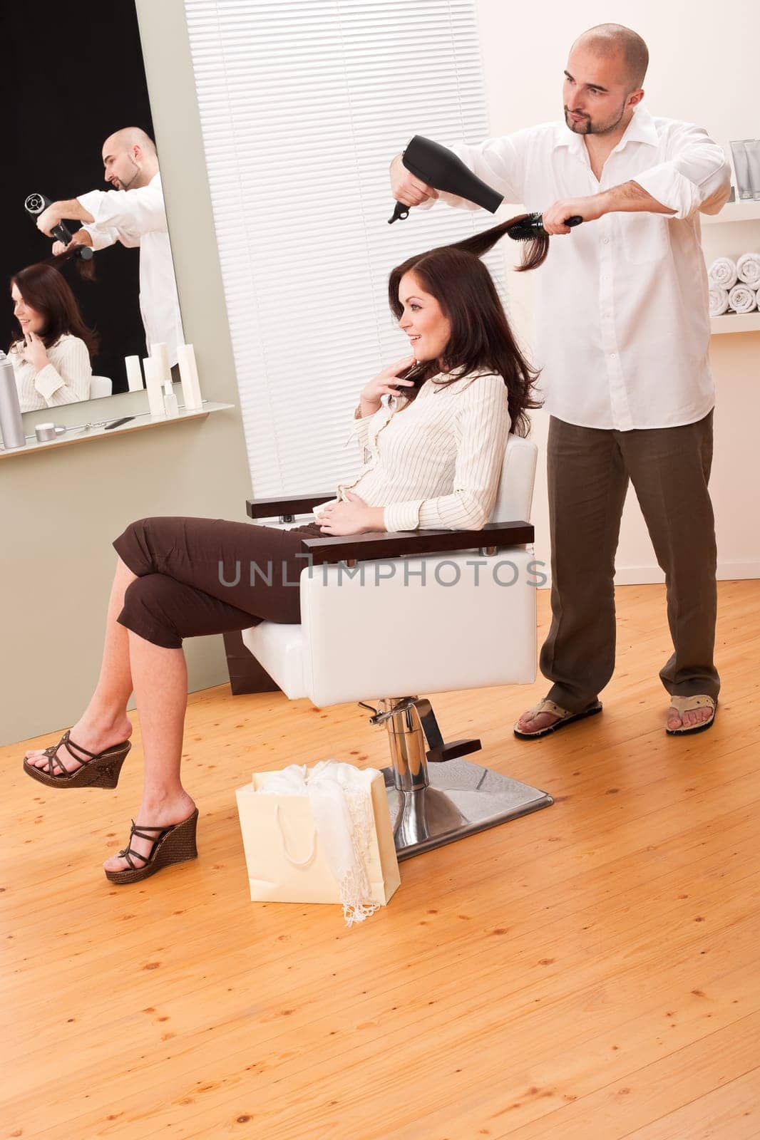 Professional male hairdresser with hair dryer and hair brush at salon with female customer, shopping bag on floor