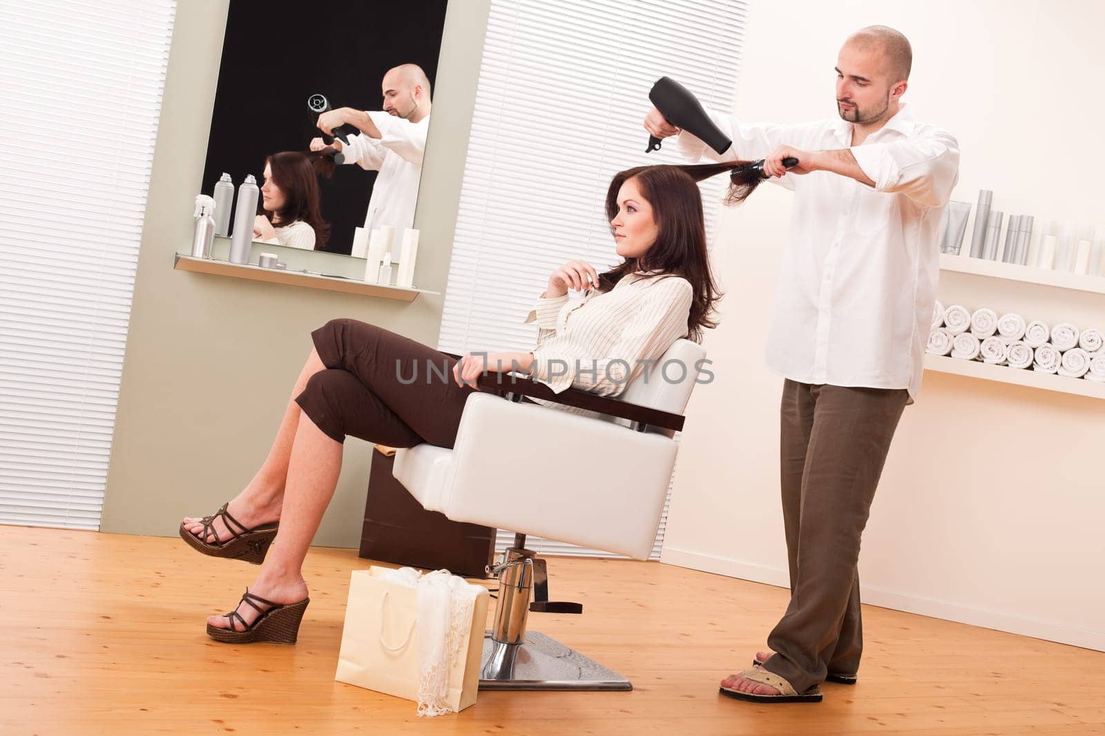 Professional male hairdresser with hair dryer and hair brush at salon with female customer, shopping bag on floor