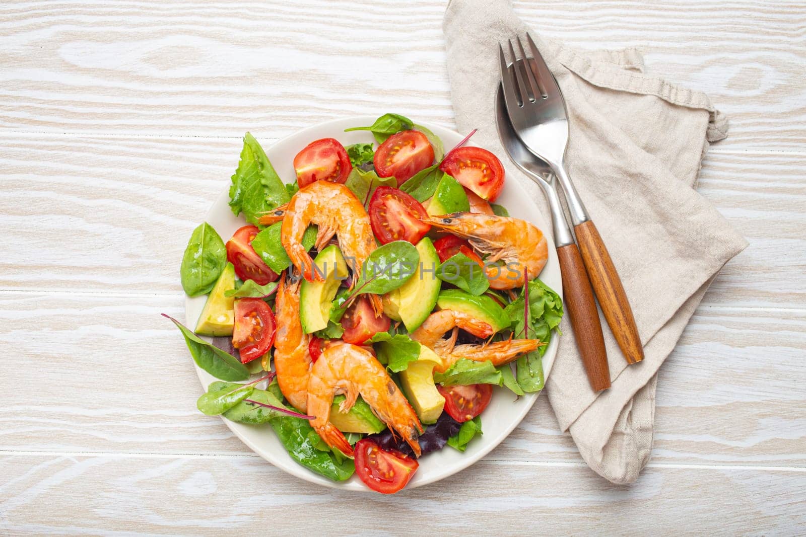 Healthy salad with grilled shrimps, avocado, cherry tomatoes and green leaves on white plate with cutlery on white wooden rustic background top view. Clean eating, nutrition and dieting concept..