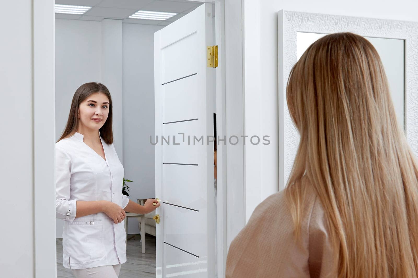 Young female cosmetologist confidently steps into a room where a patient sits in a robe