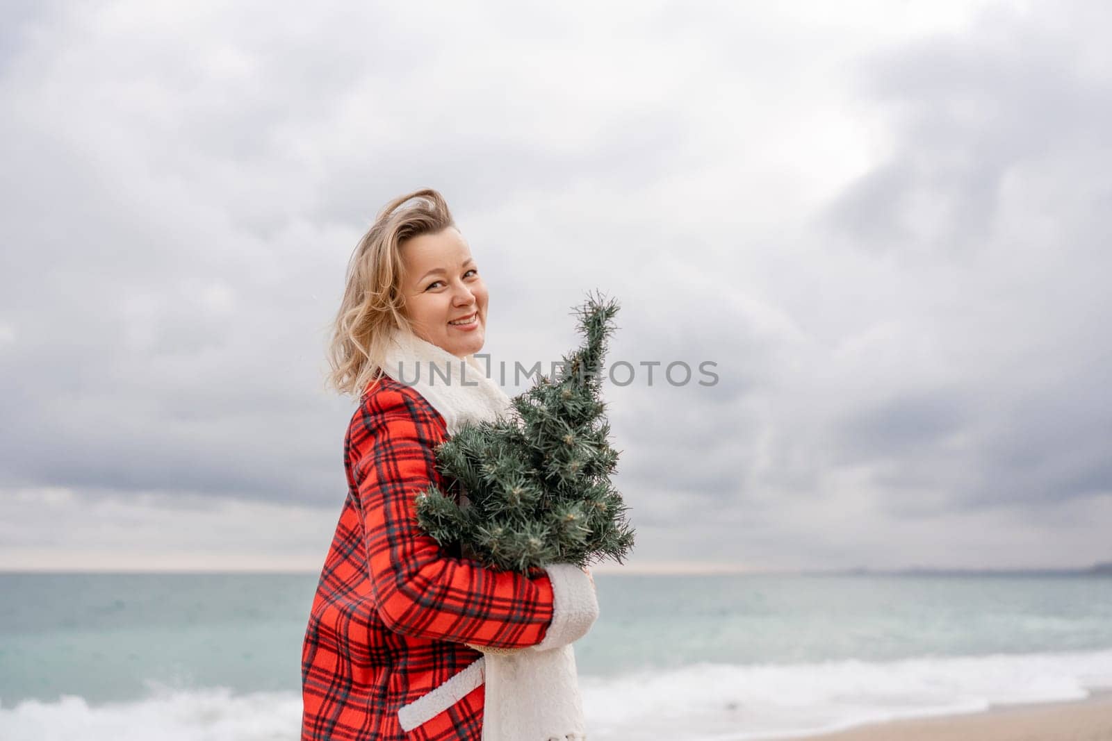 Blond woman holding Christmas tree by the sea. Christmas portrait of a happy woman walking along the beach and holding a Christmas tree in her hands. Dressed in a red coat, white dress