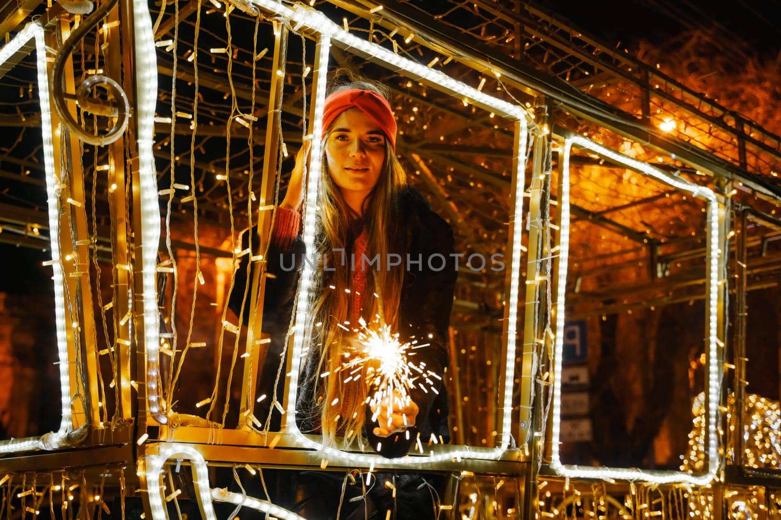 Woman holding sparkler night while celebrating Christmas outside. Dressed in a fur coat and a red headband. Blurred christmas decorations in the background. Selective focus by Matiunina