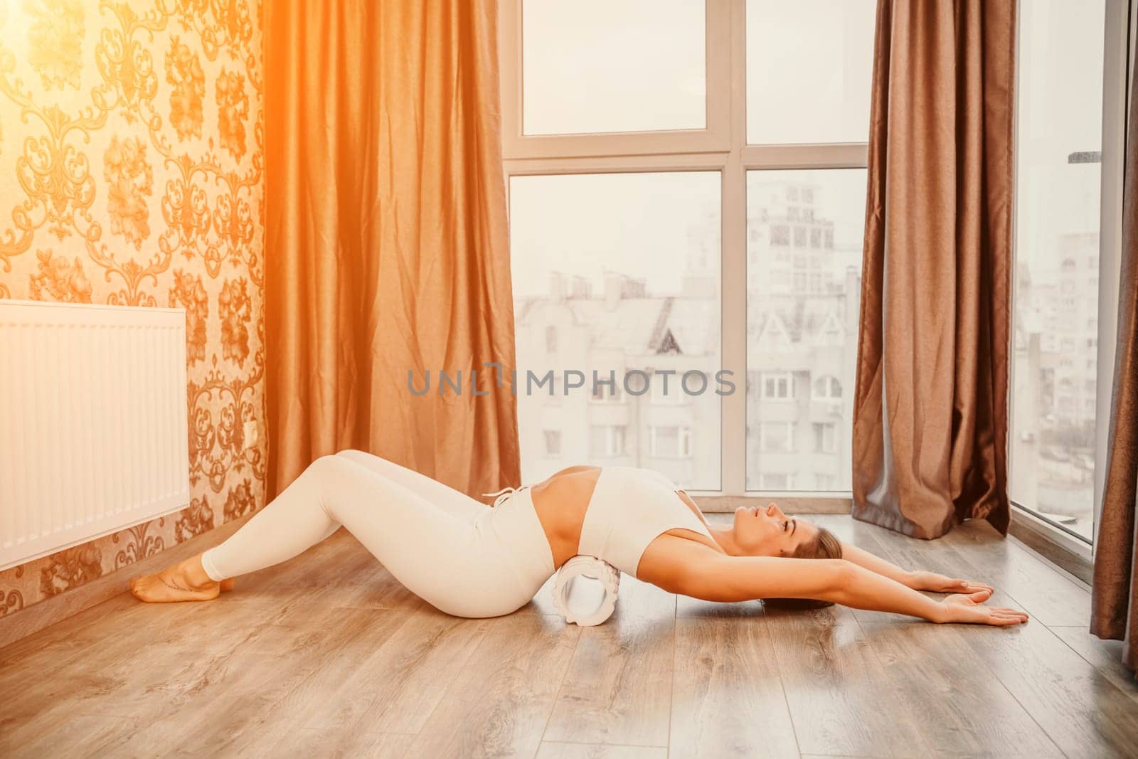 Adult athletic woman, in white bodysuit, performing fascia exercises on the floor - caucasian woman using a massage foam roller - a tool to relieve tension in the back and relieve muscle pain - the concept of physiotherapy and stretching training by Matiunina