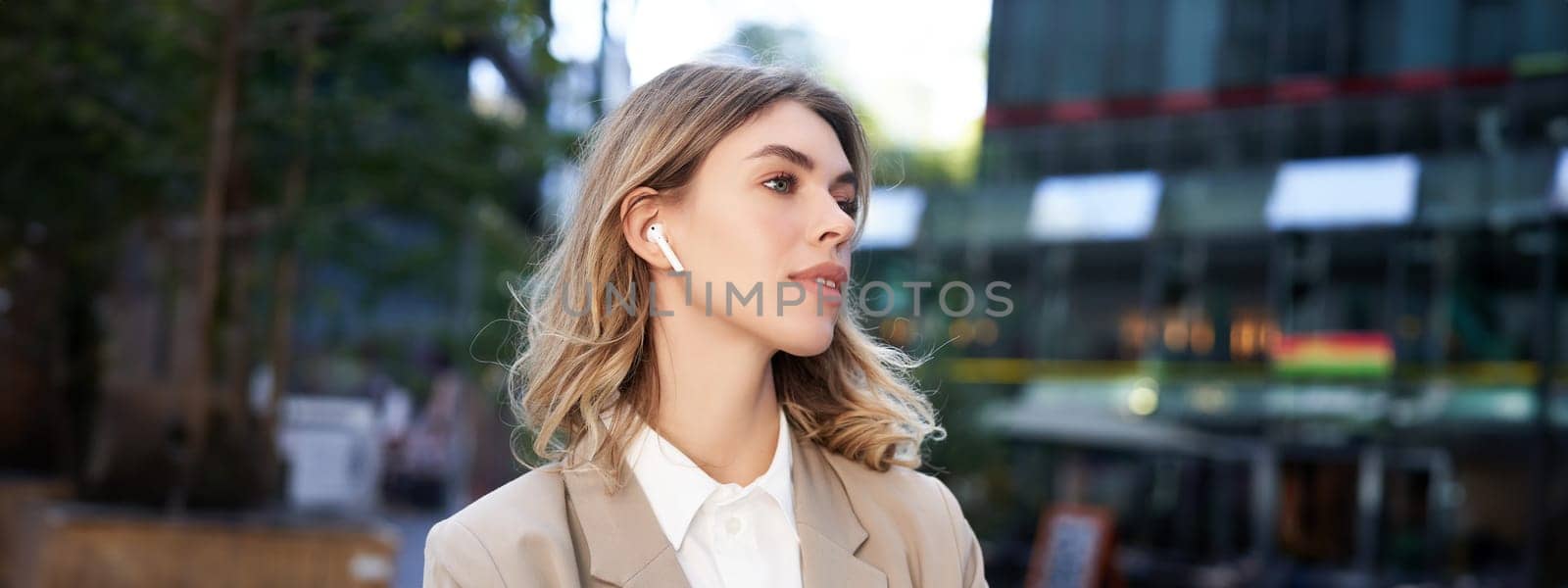 Close up portrait of blond businesswoman, confident corporate woman in headphones and beige suit, posing outdoors on street of city center by Benzoix