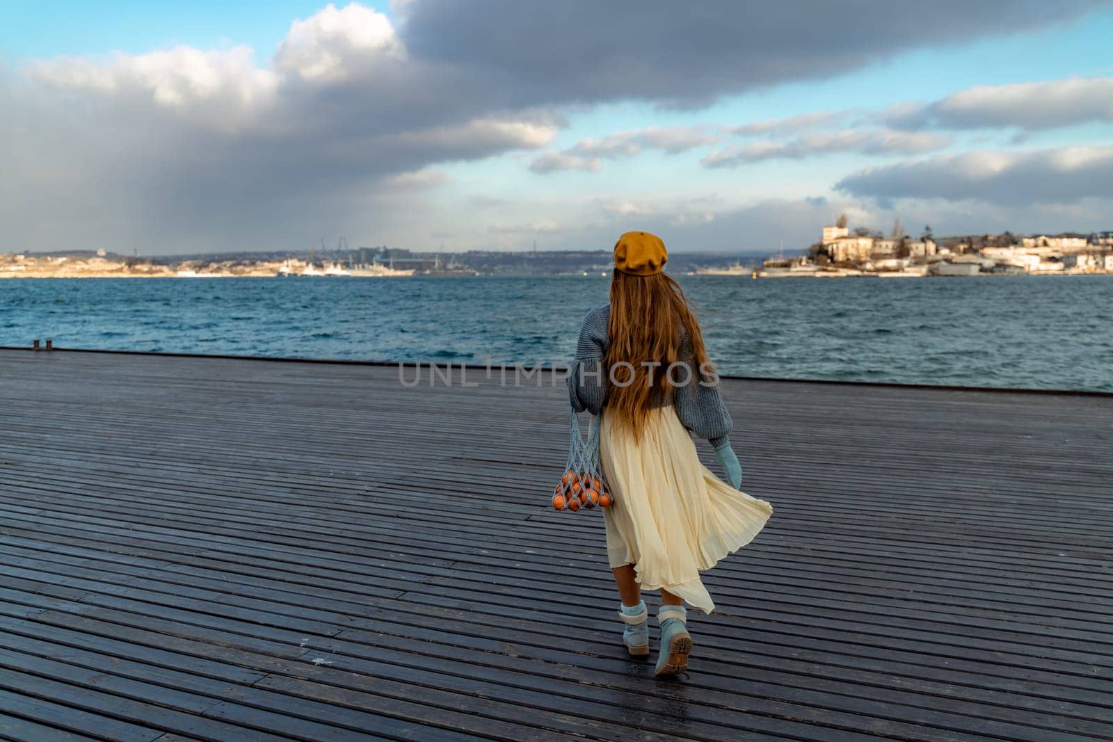 Outdoors fashion portrait of a beautiful middle aged woman walking on the beach. Marine background. Dressed in a stylish warm blue sweater, yellow skirt and beret. by Matiunina