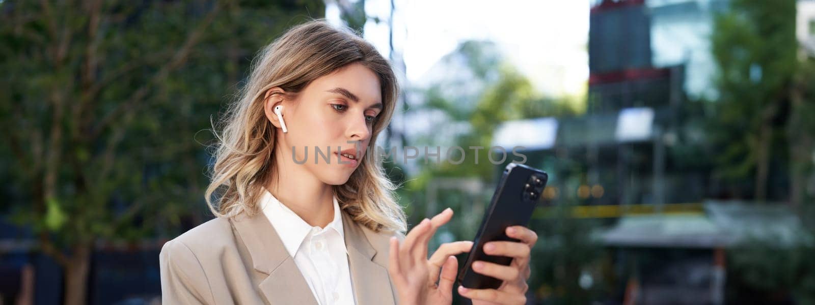 Successful businesswoman in beige suit, wireless headphones, looking at mobile phone, using smartphone app, standing outdoors on street.