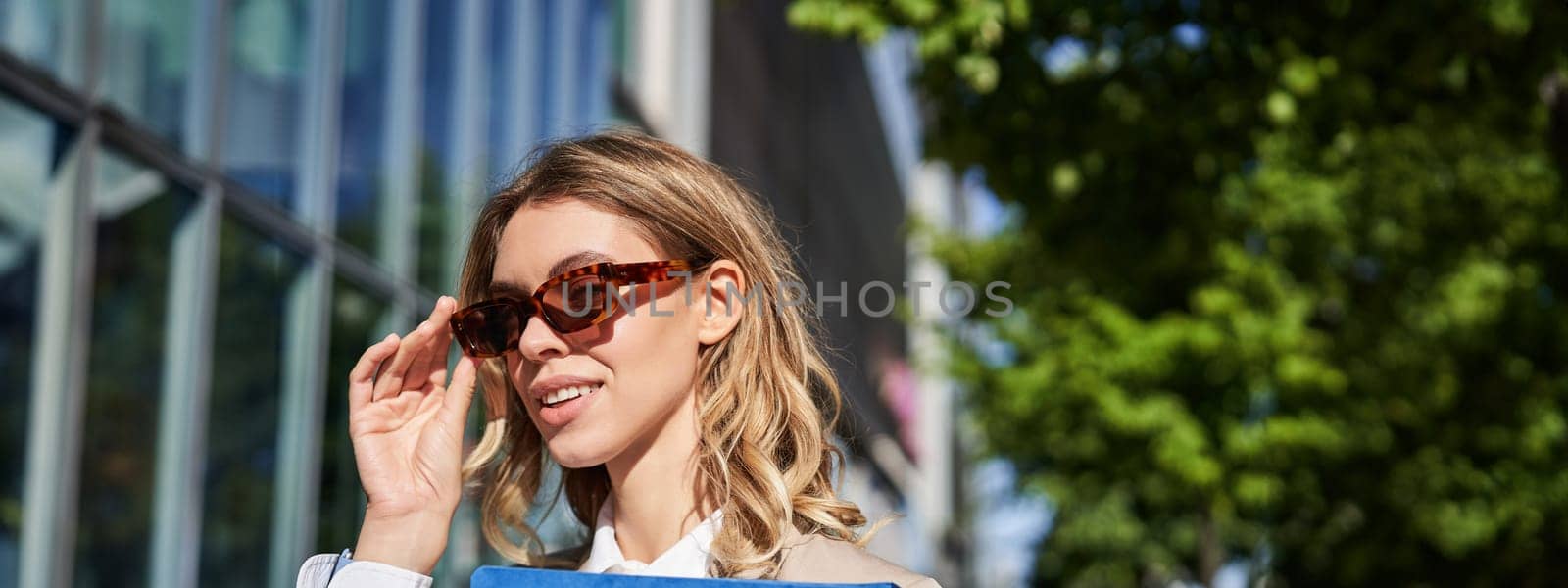 Close up portrait of smiling young woman in sunglasses, corporate suit, holding a folder with work documents, going to an interview, standing outdoors.