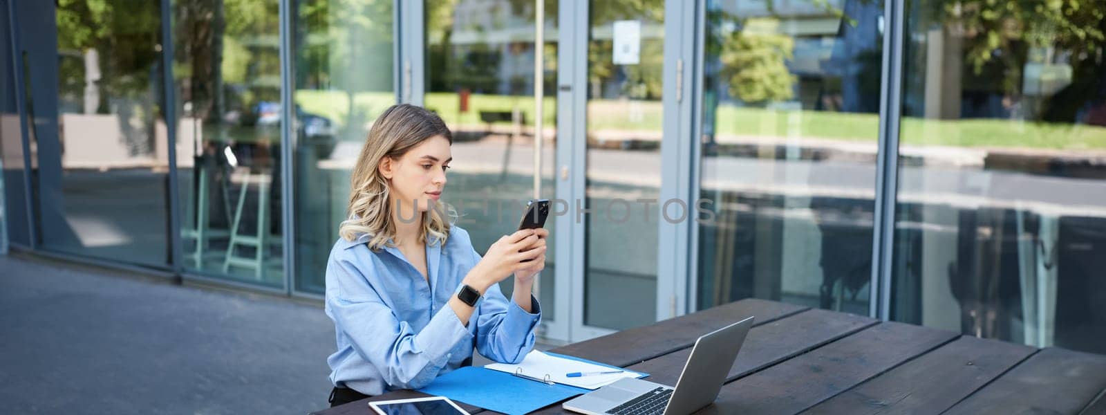 Portrait of businesswoman sitting outdoors and working. Young corporate woman looking at her smartphone, sitting outside with laptop and digital tablet.