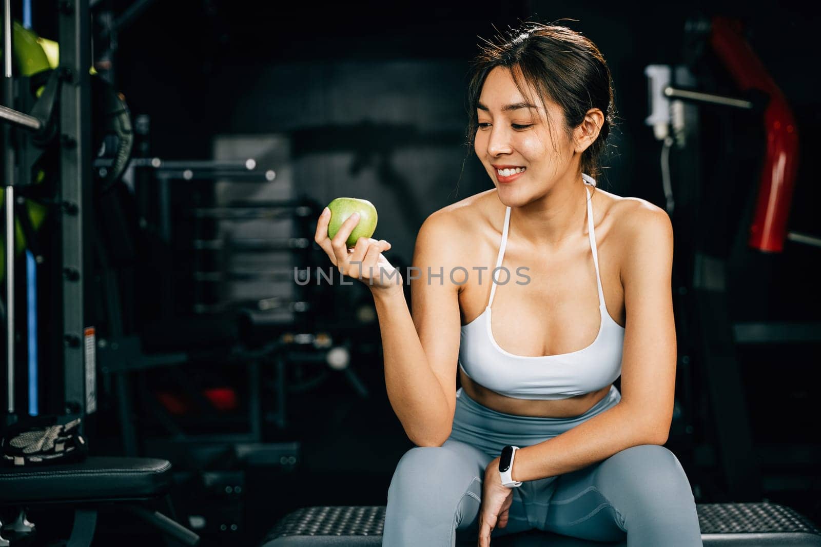 A young woman holds a green apple in a gym, reminding viewers of the importance of a balanced diet for achieving fitness goals. Healthy fitness and eating lifestyle concept