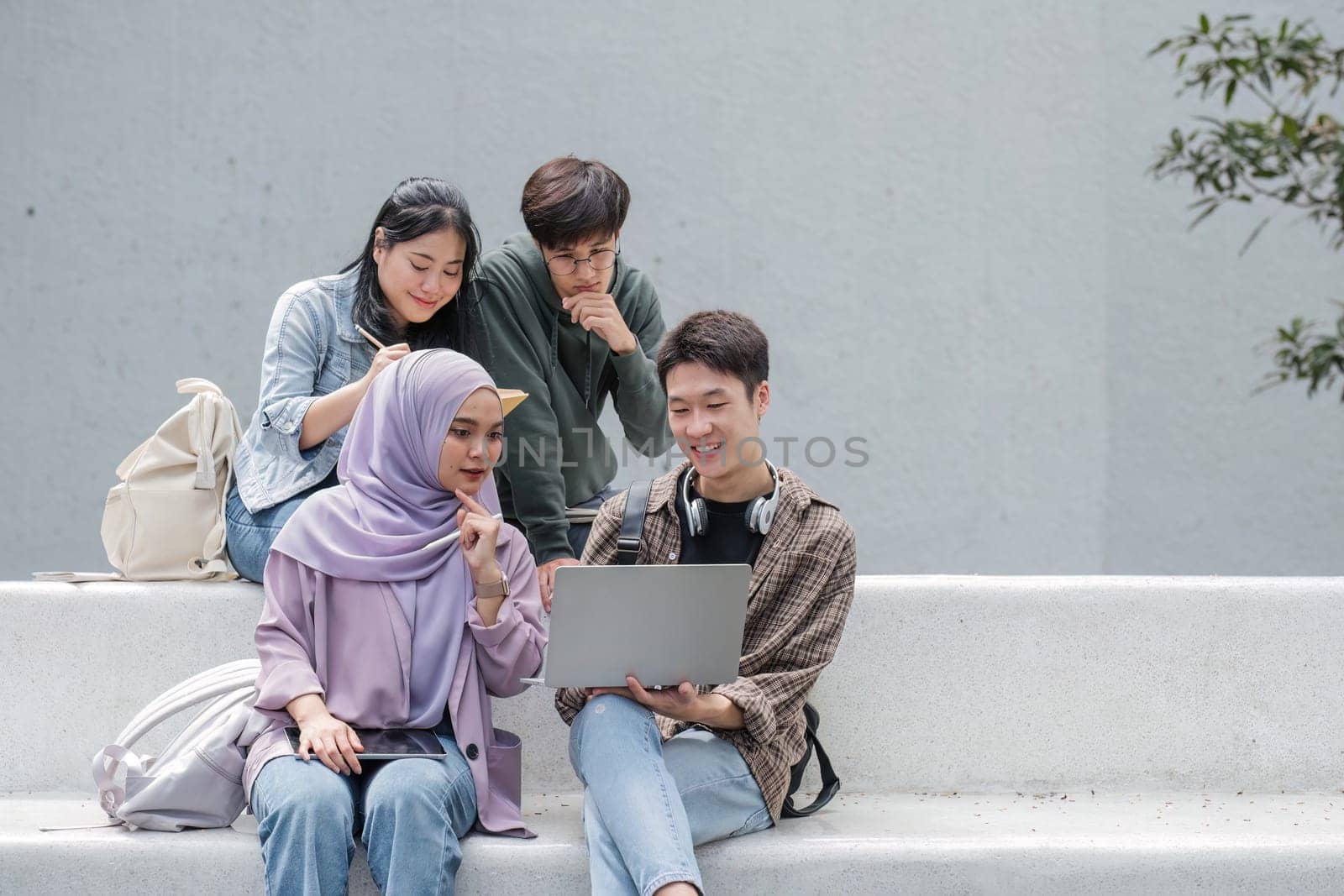 Group of happy young Asian college students sitting on a bench looking at a laptop screen, discussing and brainstorming on their school project together by wichayada