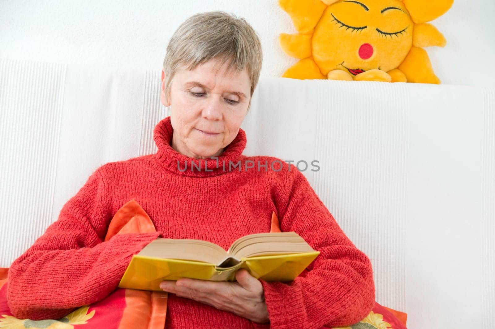 mature woman in red sweater,short and gray hair sitting on a white couch and reading a yellow book