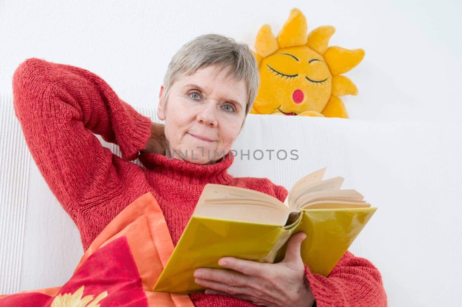 mature woman in red sweater,short and gray hair sitting on a white couch and reading a yellow book