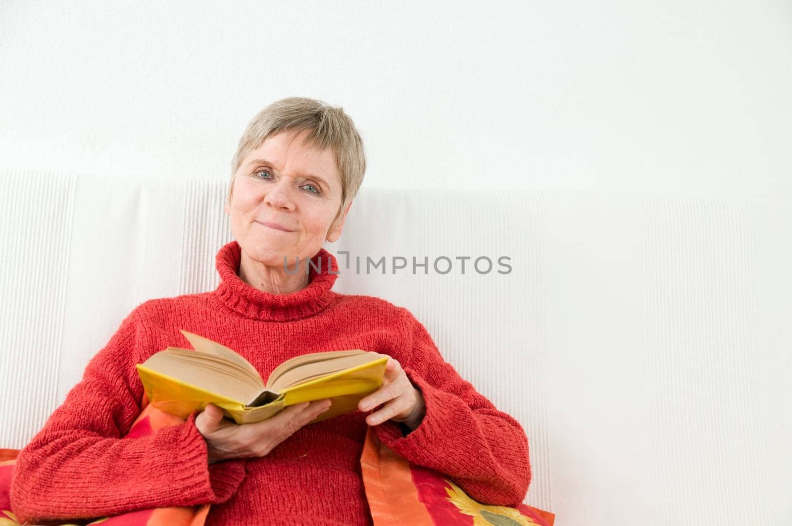 mature woman in red sweater,short and gray hair sitting on a white couch and reading a yellow book
