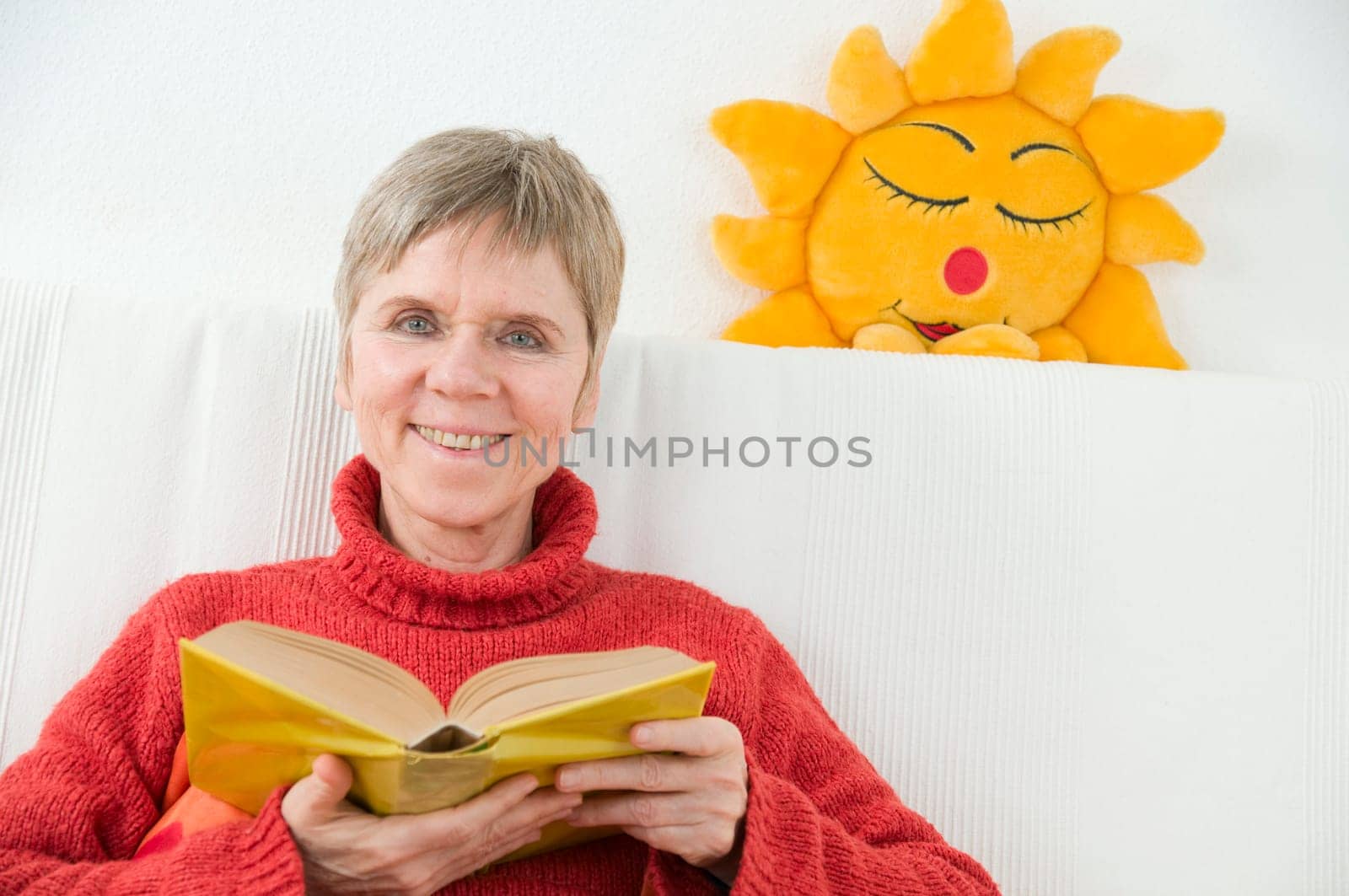 mature woman in red sweater,short and gray hair sitting on a white couch and reading a yellow book