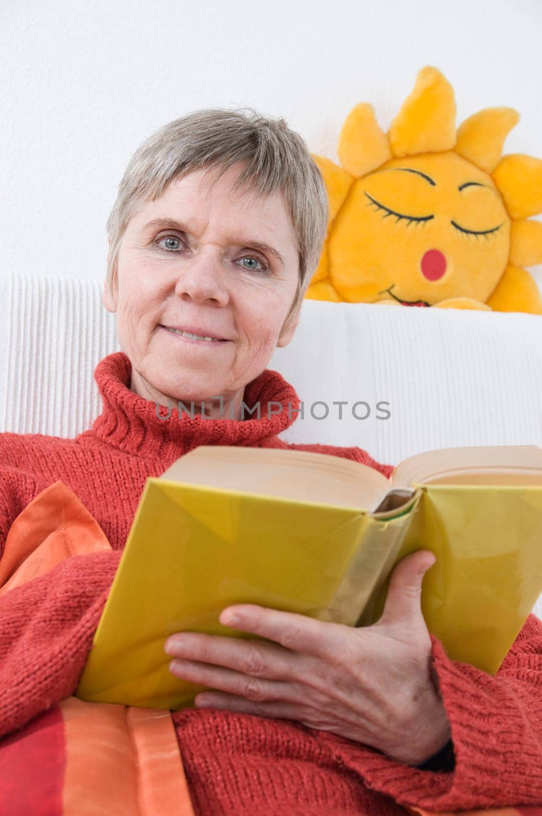 mature woman in red sweater,short and gray hair sitting on a white couch and reading a yellow book