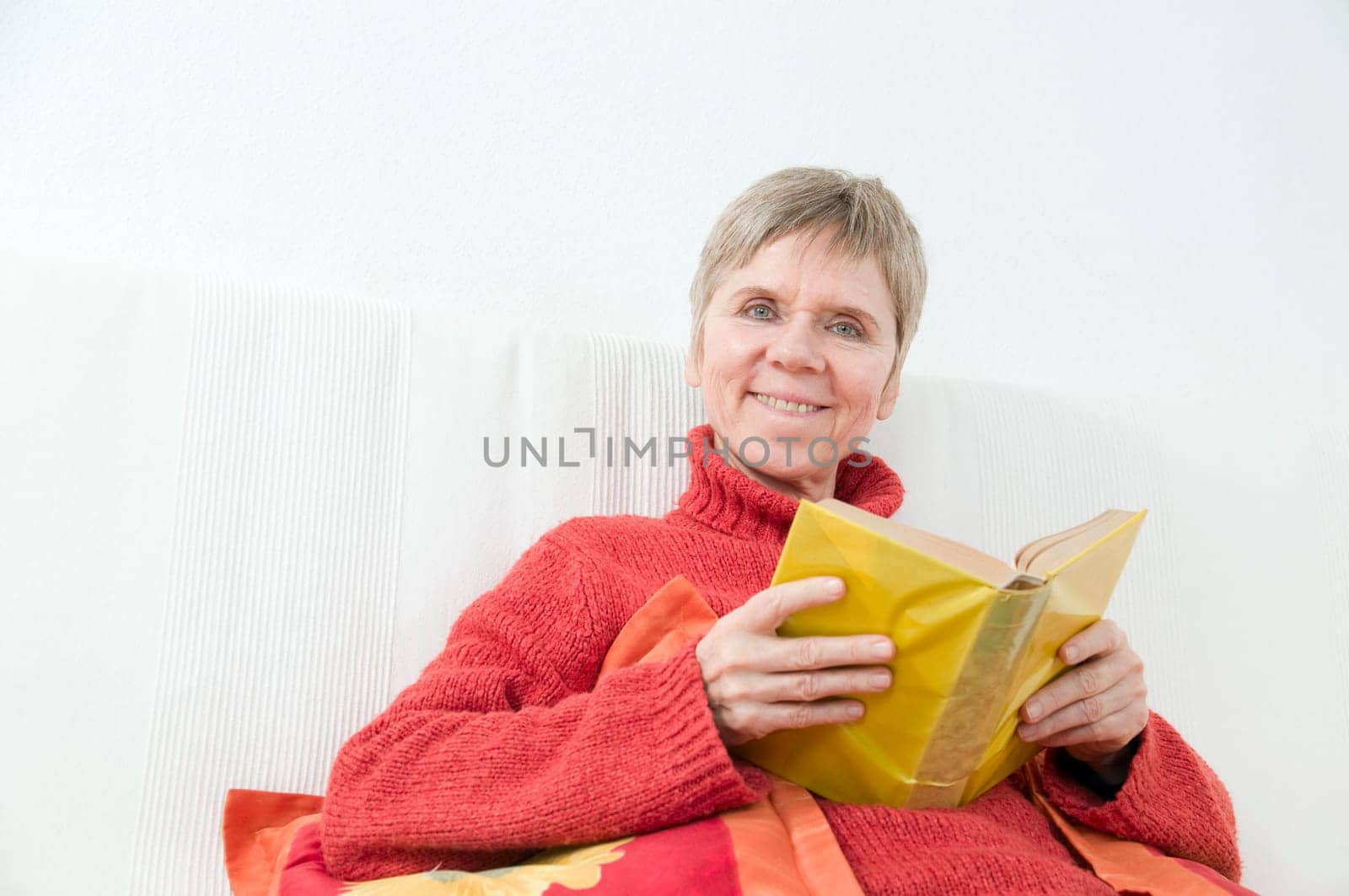 mature woman in red sweater,short and gray hair sitting on a white couch and reading a yellow book
