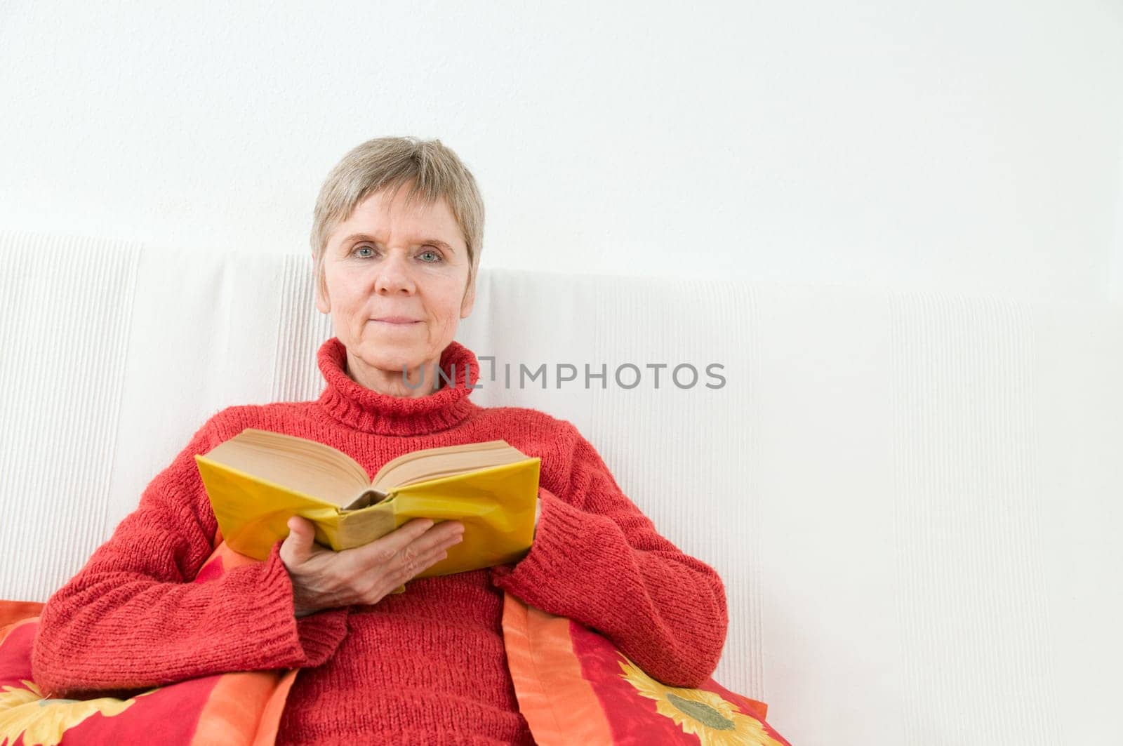 mature woman in red sweater,short and gray hair sitting on a white couch and reading a yellow book