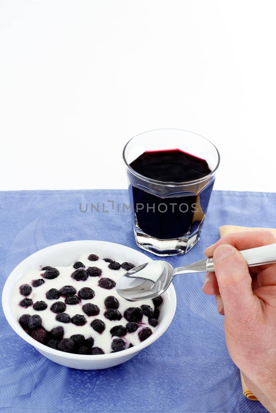 Hand with spoon about to eat a bowl of thawing frozen blueberries with vanilla yogurt, glass of organic blueberry juice.