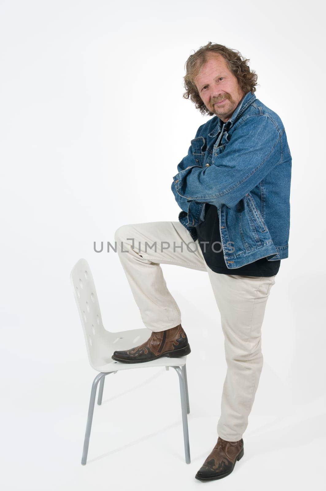 Elderly man in white pants and blue denim jacket puts one leg on the seat of a white chair
