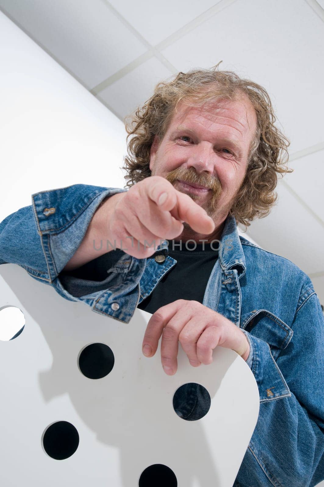 mature man sitting astride a chair pointing with index finger towards camera