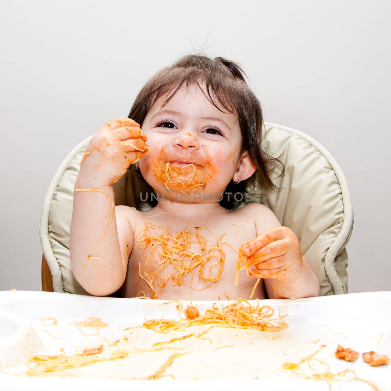 Happy smiling baby having fun eating messy covered in Spaghetti Angel Hair Pasta red marinara tomato sauce.