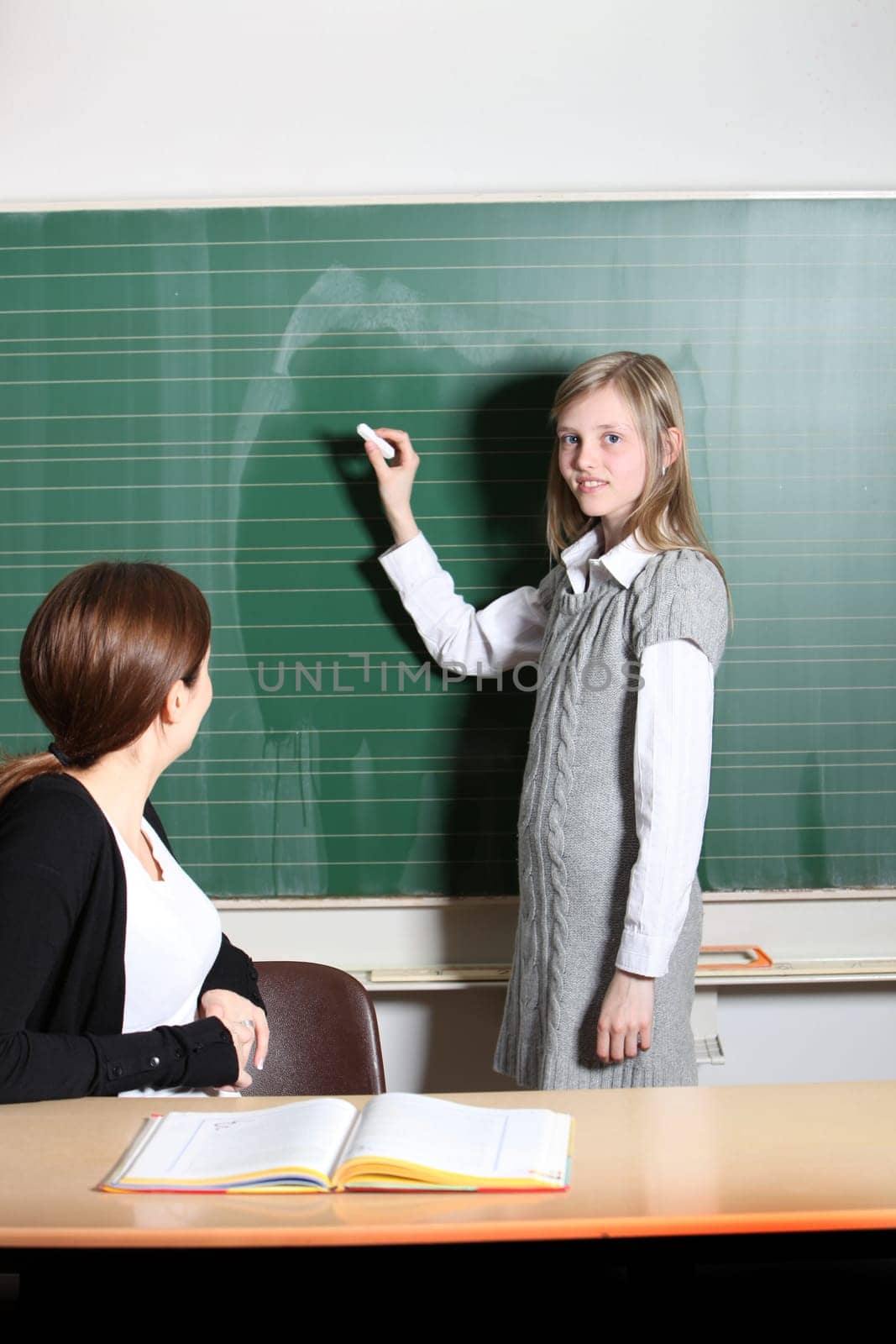 Schoolgirl in the classroom writes on the blackboard. The teacher sits at the desk and watches