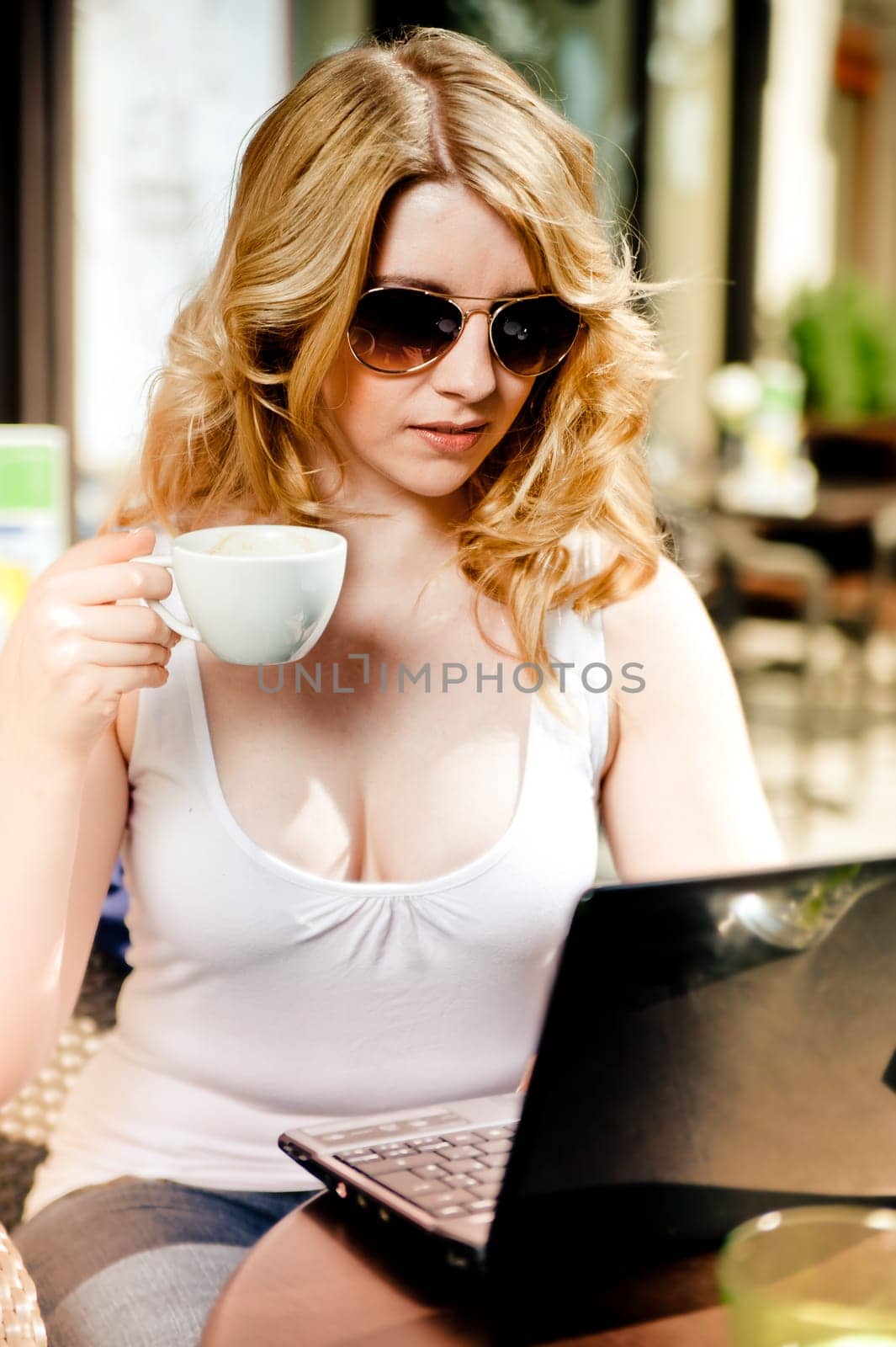Blonde Woman sitting in a Cafe, looking into her Laptop with Cup in her Hand