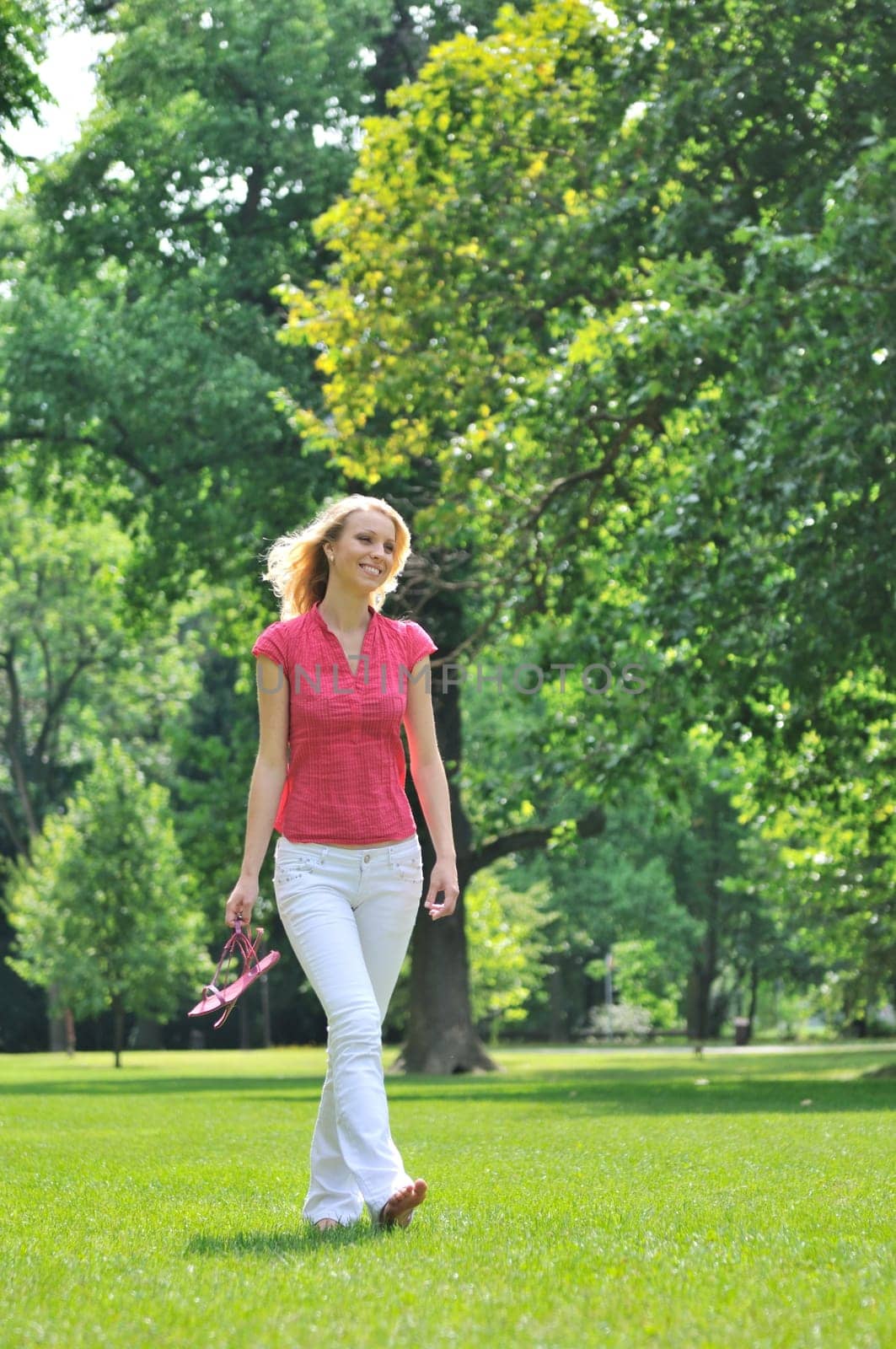 Smiling young woman holding her shoes and walking barefoot on grass in park