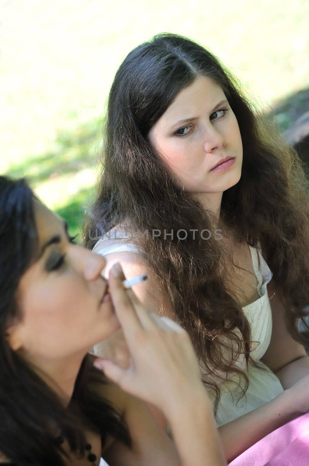 Youth culture - two young people outdoors, one woman smoking cigarette annoys and iritates another girl