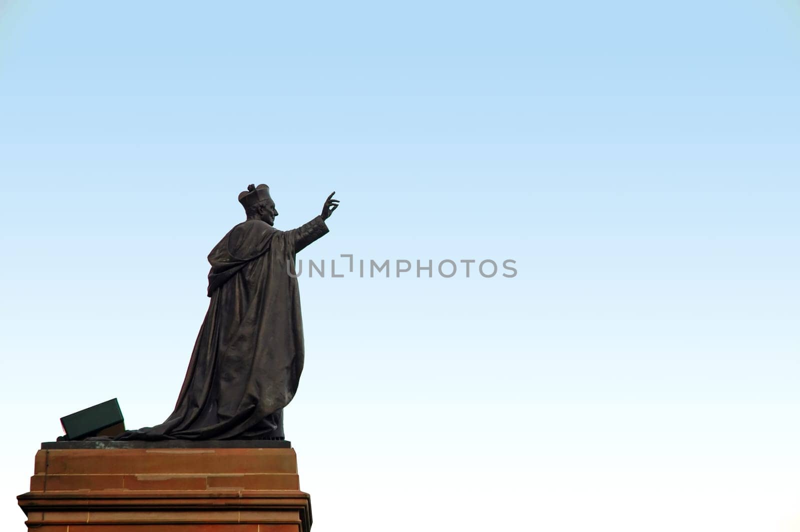dark grey pope sculpture with one hand lifted up, sculpture is on brown stand, light blue sky,
