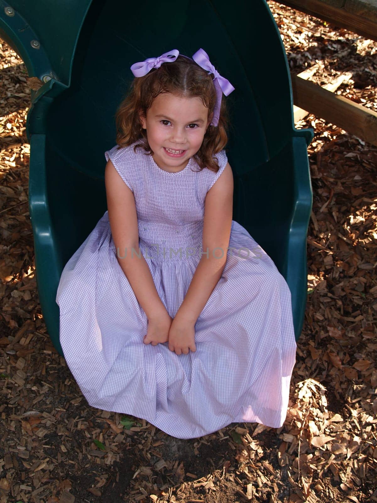 Young Girl Sitting at the end of a green tube slide on a playground.