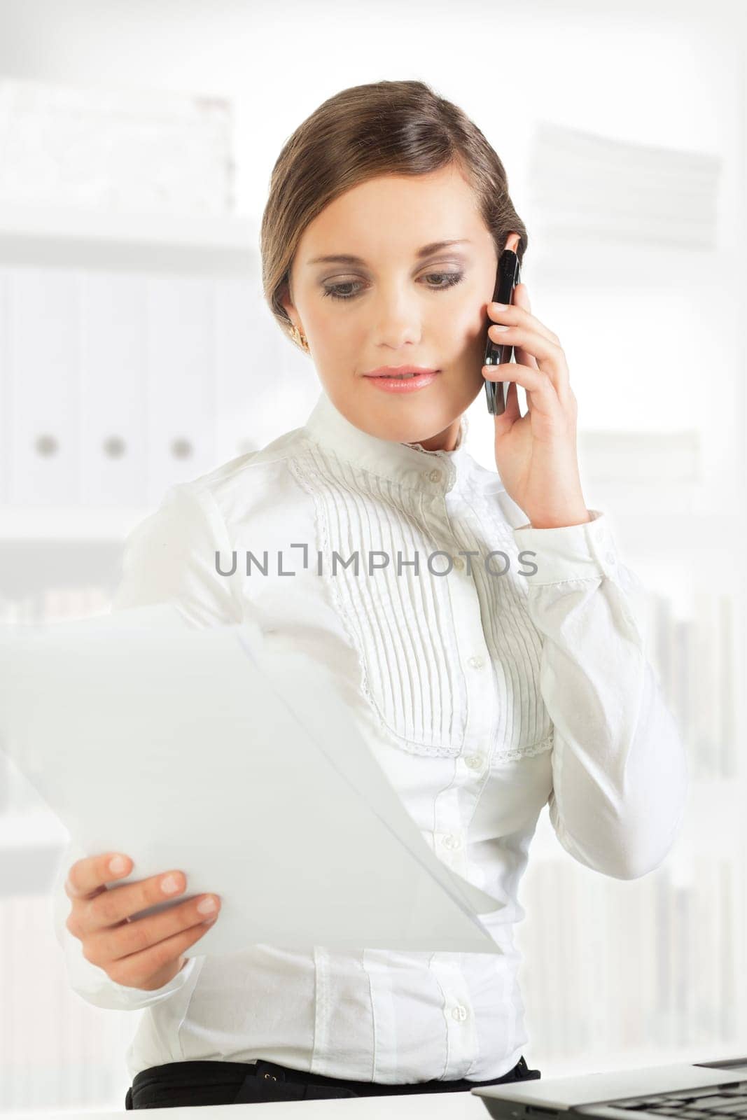 Pretty young businesswoman reading papers while speaking on the phone