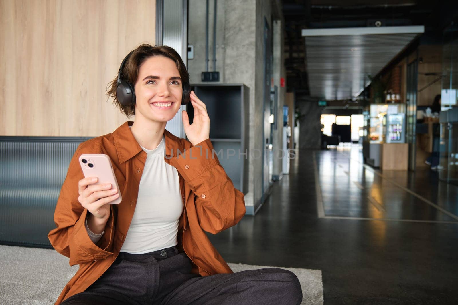 Portrait of happy young employee, woman manager sitting on floor, listening music in wireless headphones, holding mobile phone, relaxing in lounge room. People and lifestyle concept