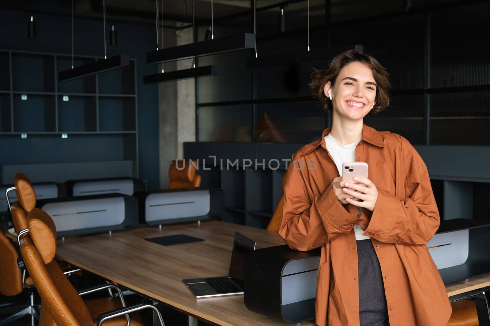 Young modern woman, office employee standing in conference room with smartphone and wireless headphones.
